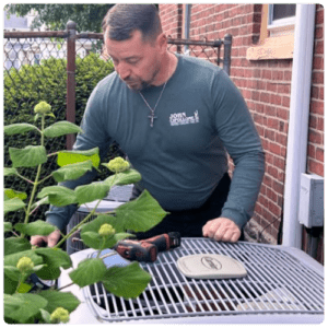 Man in a teal long-sleeve shirt leaning over a grill on a brick patio, with a drill and a stone on the grate in front of him.