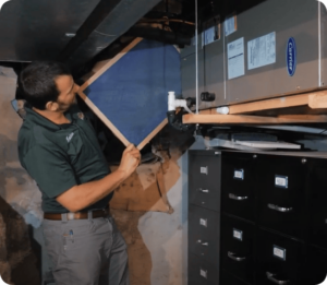 HVAC technician in a green shirt installs a blue air filter frame on an HVAC unit in a basement.